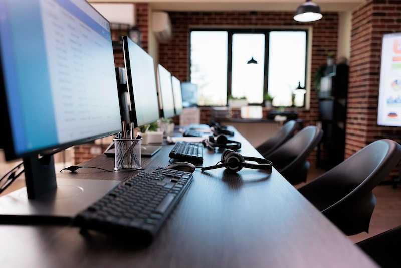 An empty call center workstation showing multiple monitors, keyboards, and headsets arranged on a dark desk, with exposed brick walls and windows in the background.